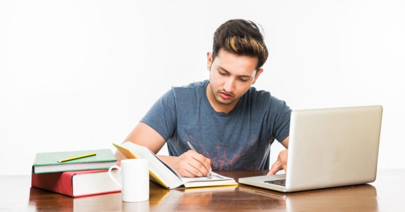 student studying on laptop and from books kept over table with coffee mug, over plain white background.