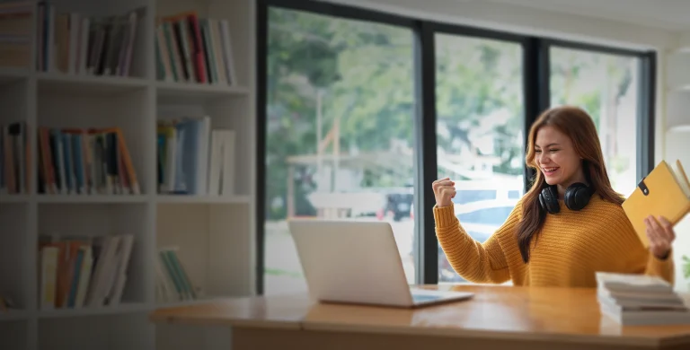 A happy student in a yellow sweater celebrating a good Digital SAT score report which is on her laptop computer