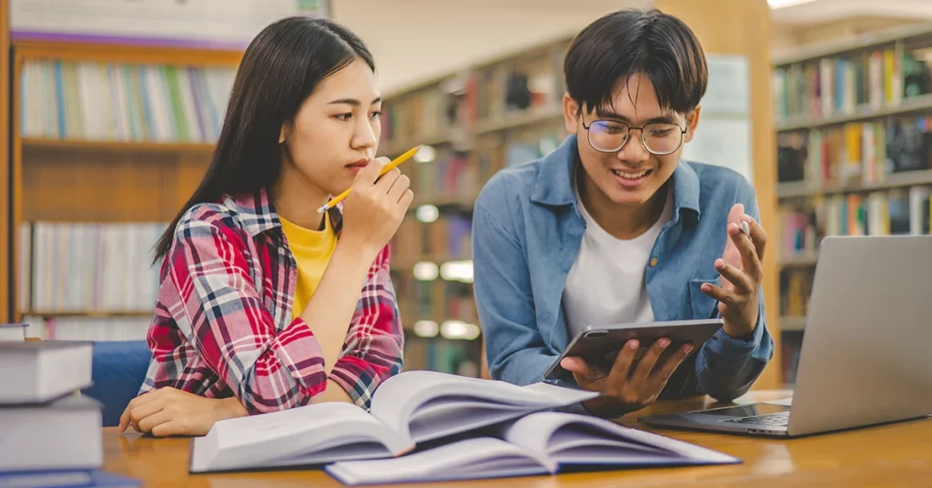 Two students studying together with books, tablet, and laptop in a library.