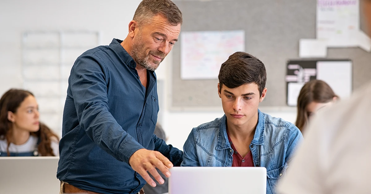 Teacher assisting a high school student with laptop during a classroom tutoring session.