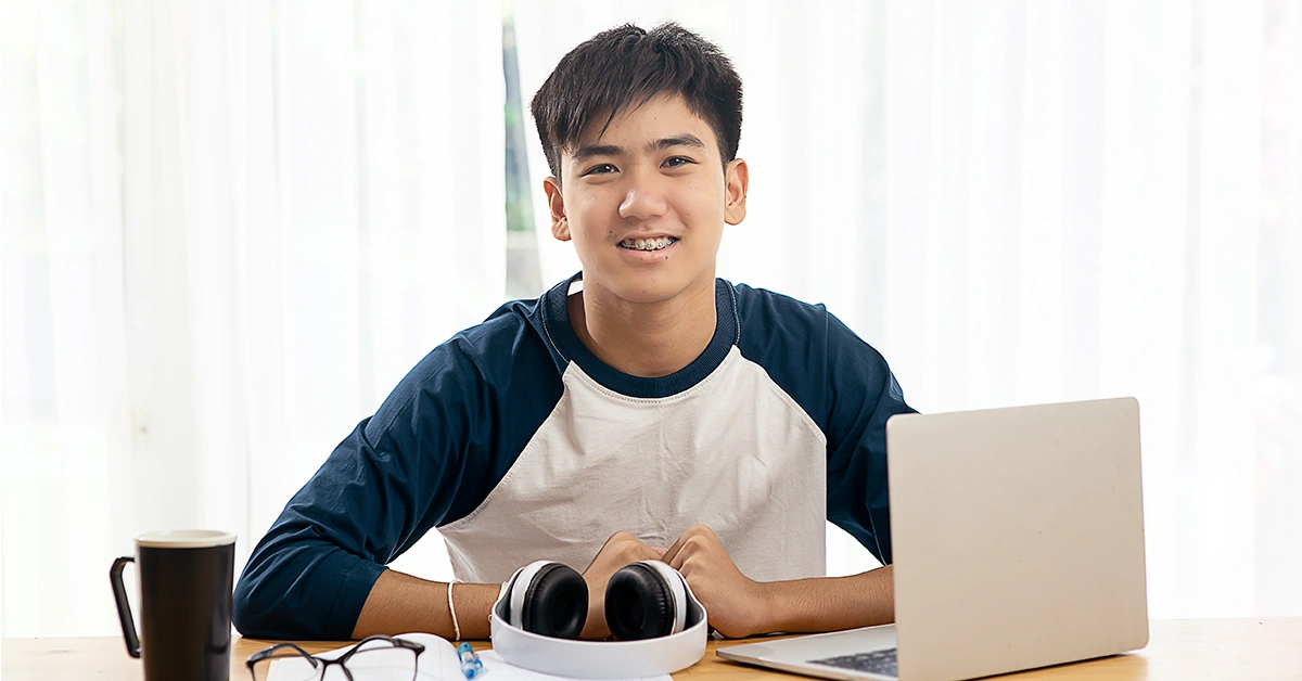 Smiling high school student studying at a desk with a laptop and headphones, staying motivated while preparing for the SAT at home