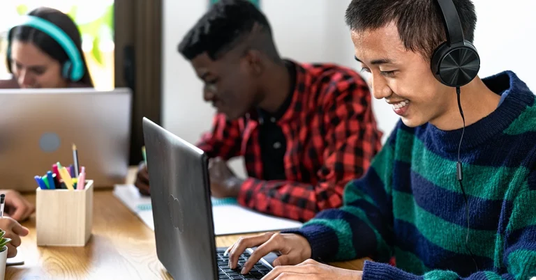 High school students wearing headphones studying with laptops, smiling and engaged in learning, possibly listening to SAT prep podcasts.